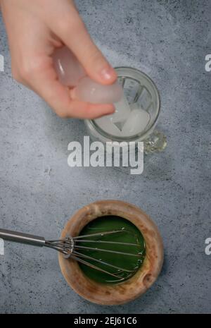 Image verticale de l'argile bol matcha thé tandis que la main fille met des cubes de glace dans le verre de cristal sur fond de pierre grise. Thé de matcha japonais 2021. Banque D'Images