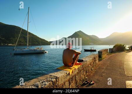 PERAST, MONTÉNÉGRO - 14 JUILLET 2016 : un homme en maillot de bain et une casquette rouge prend un bain de soleil au coucher du soleil le long de la route qui traverse le village de Perast Banque D'Images