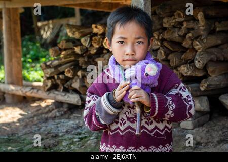 Portrait d'un petit garçon asiatique tenant son jouet pourpre à l'extérieur de sa maison dans une zone rurale avec du bois de chauffage en arrière-plan. Banque D'Images