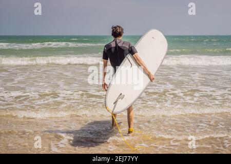 Jeune surfeur debout contre l'océan regardant les vagues, surfeur professionnel en combinaison noire tenant avec une main grande planche à voile ensoleillée Banque D'Images