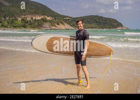 Jeune surfeur debout contre l'océan regardant les vagues, surfeur professionnel en combinaison noire tenant avec une main grande planche à voile ensoleillée Banque D'Images