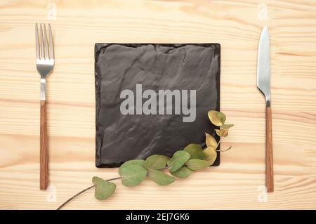 Couteau et fourchette avec plaque carrée en ardoise noire pour le texte du menu ou de la recette et branche d'eucalyptus d'or sur table en bois. Maquette planche vierge et couverts sur la lune Banque D'Images