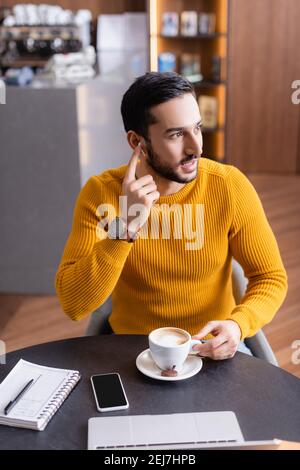 jeune indépendant arabe qui fixe des écouteurs tout en étant assis près d'une tasse de café et ordinateur portable Banque D'Images