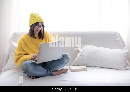 Élégante et gaie brune fille dans des verres et un chapeau assis sur le canapé à la maison et travaille à un ordinateur portable copie espace. Banque D'Images