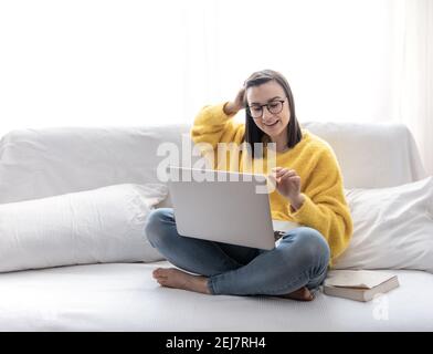 Une fille brune élégante dans un chandail jaune s'assoit à la maison sur le canapé dans une pièce lumineuse et travaille à distance sur un ordinateur portable. Banque D'Images