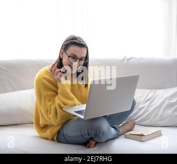 Une fille brune élégante dans un chandail jaune est assise avec un ordinateur portable à la maison sur le canapé et sourit. Banque D'Images