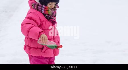 Une jolie fille avec une bouteille de peinture dessine sur la neige. Le concept de la créativité des enfants et du plaisir d'hiver. Banque D'Images