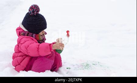 Une jolie fille avec une bouteille de peinture dessine sur la neige. Le concept de la créativité des enfants et du plaisir d'hiver. Banque D'Images