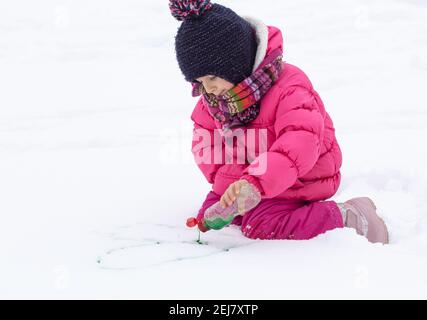 Une jolie fille avec une bouteille de peinture dessine sur la neige. Le concept de la créativité des enfants et du plaisir d'hiver. Banque D'Images