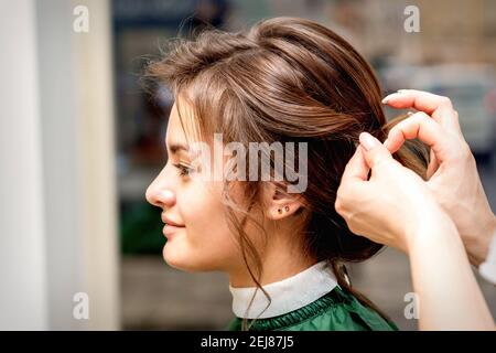 Vue latérale des mains du coiffeur styliste coiffant les cheveux la belle jeune femme brune caucasienne dans un salon de beauté Banque D'Images