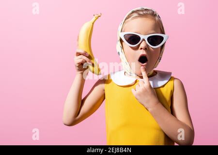 fille choquée dans le foulard et lunettes de soleil tenant la banane isolée sur rose Banque D'Images