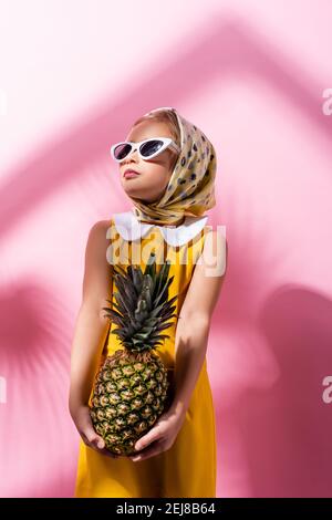 élégante fille en foulard et lunettes de soleil tenant l'ananas sur le rose Banque D'Images