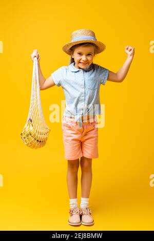 toute la longueur de l'enfant excité dans un chapeau de paille, réutilisable sac à ficelle avec bananes sur jaune Banque D'Images