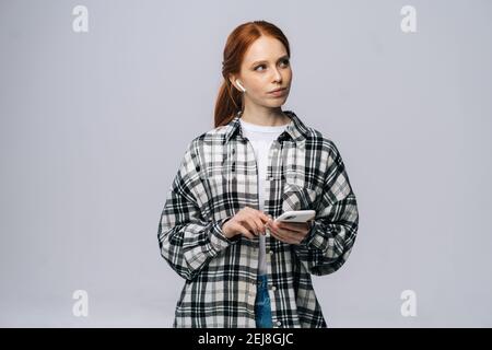 Penser jeune femme portant des écouteurs sans fil taper un message en ligne sur un fond blanc isolé. Banque D'Images