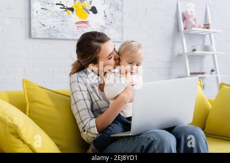 un jeune freelance embrasse un petit enfant assis sur un canapé jaune avec ordinateur portable Banque D'Images