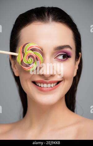 jeune femme heureuse avec des ombres à paupières roses couvrant l'œil avec lollipop isolé sur gris Banque D'Images