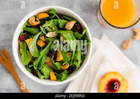 Salade d'arugula, de pêches jaunes, de noix de cajou et de canneberges séchées dans un bol blanc avec une fourchette en bois, un verre de jus d'orange et une moitié de pêche. Banque D'Images