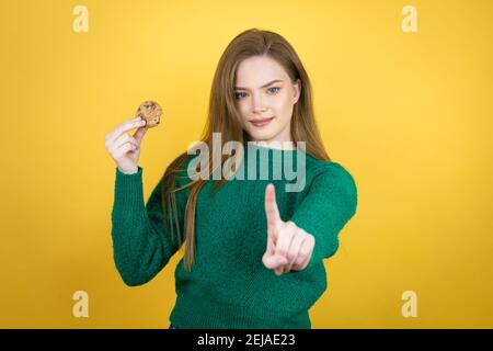 Jeune belle femme mangeant le cookie de chocolat sur fond jaune montrant et de pointer vers le haut avec les doigts numéro un pendant est grave Banque D'Images