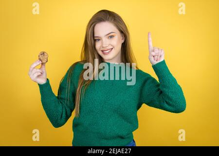 Jeune belle femme mangeant le cookie de chocolat sur fond jaune montrant et en pointant vers le haut avec les doigts numéro un tout en souriant confiant et heureux Banque D'Images