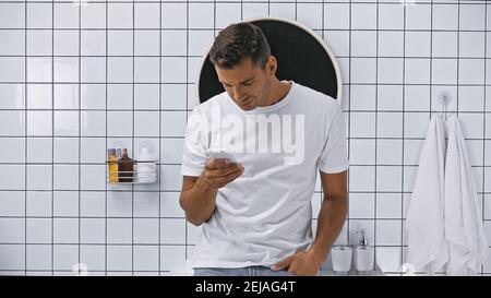 jeune homme en t-shirt blanc sur téléphone mobile dans salle de bains Banque D'Images