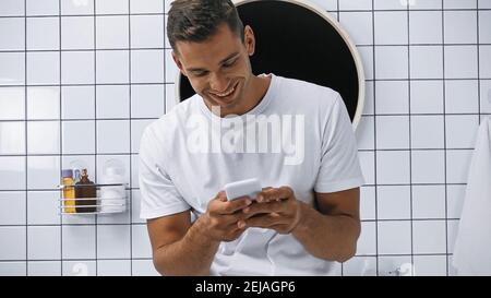 homme souriant dans un t-shirt blanc discutant sur un smartphone dans la salle de bains Banque D'Images