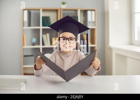 Petite fille avec des lunettes est assise à la table dans un chapeau de diplômé et avec un diplôme en mains. Banque D'Images