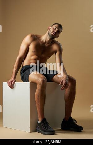 Portrait complet d'un homme fort et beau avec corps musculaire assis sur une chaise blanche et regardant la caméra en studio Banque D'Images