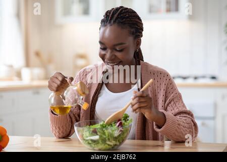 Femme afro-américaine souriante cuisant de la salade de légumes dans la cuisine, ajoutant de l'huile d'olive Banque D'Images