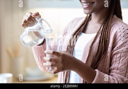 Souriante femme afro-américaine versant de l'eau de la carafe au verre, gros plan Banque D'Images