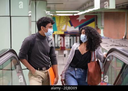 Jeune couple latin portant un masque de protection se promenant dans les escaliers mécaniques de la gare ou de la station de métro. Nouvelle norme dans les transports publics. Banque D'Images