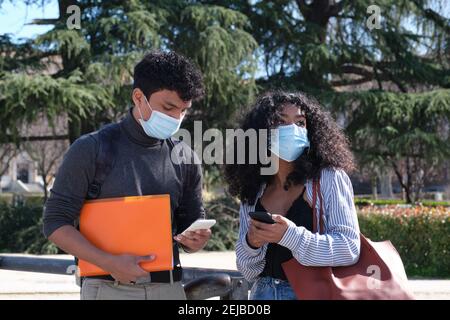 Jeune couple latin portant un masque de protection regardant le smartphone. Nouvelle norme sur le campus universitaire. Banque D'Images