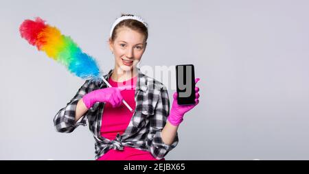 Bannière. Jolie fille souriante vêtue de vêtements de femme de ménage, tenant une brosse à poussière. Blonde gaie femme pointant l'index vers un smartphone noir factice vide Banque D'Images