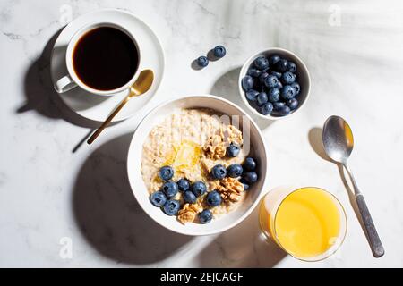 Concept petit déjeuner. Flocons d'avoine avec baies et noix, une tasse de café et un verre de jus d'orange sur fond de marbre blanc. Banque D'Images