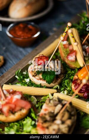 gros plan de quelques pinchos vegan espagnols, faits avec du pain avec différentes garnitures, servi comme encas ou hors-d'œuvre sur un plateau en bois, sur un bois rustique gris Banque D'Images