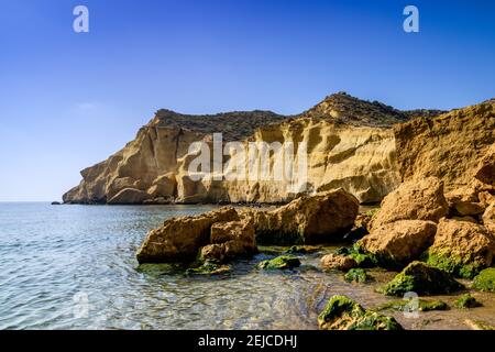 Une crique paisible et idyllique de l'océan dans la Méditerranée avec jaune falaises de grès derrière et algues vertes couvertes de rochers sur le plage de sable Banque D'Images