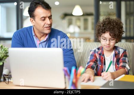 Portrait d'un père hispanique d'âge moyen passant du temps avec son fils. Petit garçon assis à la réception avec son père et faisant ses devoirs. Éducation en ligne, concept de homeschooling Banque D'Images