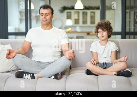 Calme-toi. Un père latin détendu d'âge moyen enseignant à son fils de méditer pendant qu'ils sont assis ensemble sur un canapé à la maison. Concept de saines habitudes de vie. Vue avant Banque D'Images