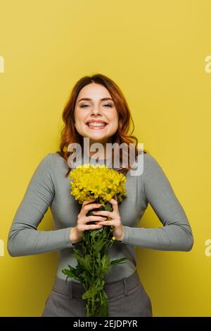 jeune femme joyeuse aux cheveux ondulés tenant des fleurs sur le jaune Banque D'Images