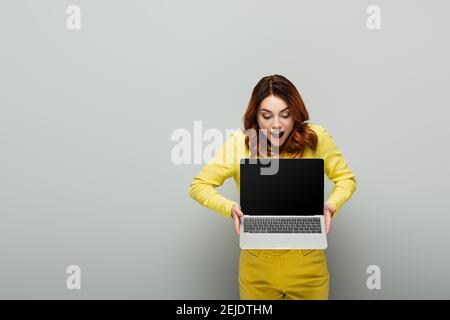 femme stupéfaite avec des cheveux bouclés tenant un ordinateur portable avec écran vierge en gris Banque D'Images