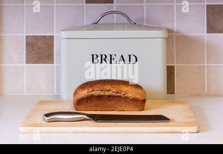 Un pain de pain complet fraîchement cuit se trouve sur un planche à pain devant une boîte de rangement avec couteau prêt à être coupé Banque D'Images