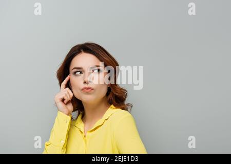 femme attentionnés avec des cheveux bouclés regardant loin tout en touchant la tête avec doigt isolé sur gris Banque D'Images