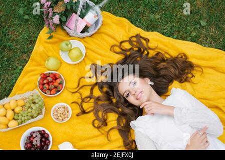 Souriante jeune fille caucasienne se trouve sur le dos à l'extérieur. Vue de dessus. Jeune femme artistique créative portant des vêtements décontractés ayant un pique-nique avec des fruits sur su Banque D'Images