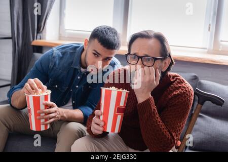 un homme inquiet tient du pop-corn tout en regardant la télévision avec son fils hispanique à la maison Banque D'Images