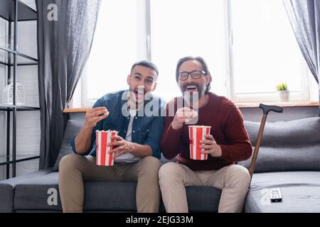 un père et un fils interraciaux excités rient tout en regardant la comédie et manger du pop-corn à la maison Banque D'Images