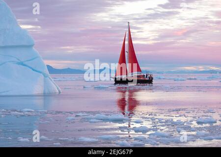 Magnifique petit voilier rouge dans l'arctique à côté d'un iceberg massif montrant l'échelle de la lumière rose de l'aube. Ilulissat, baie de Disko, Groenland. Banque D'Images