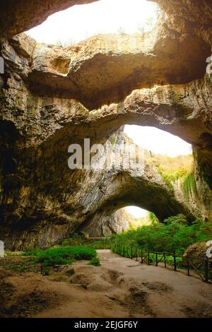 Grotte de Devetashka en Bulgarie - attraction naturelle. De hautes arches d'une immense grotte en pierre avec des trous ronds au sommet, une route touristique avec une clôture à l'intérieur du Banque D'Images