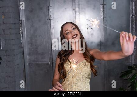 Belle fille avec de longs cheveux bouclés sourires, tient brillants sparkers dans les mains. Avec un maquillage professionnel de soirée, dans un sommet doré, contre une wa de métal gris Banque D'Images