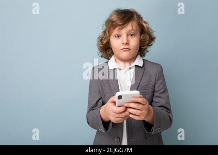 Beau garçon aux cheveux bouclés portant un costume tenant le téléphone isolé sur fond bleu, en regardant l'appareil photo Banque D'Images