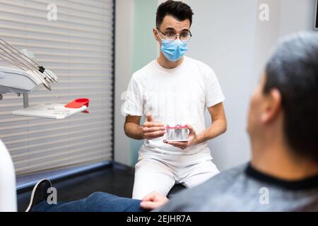 Le chirurgien dentaire avec la couronne dentaire dans les mains modèle parle au patient au sujet des prothèses. Homme au rendez-vous du dentiste Banque D'Images