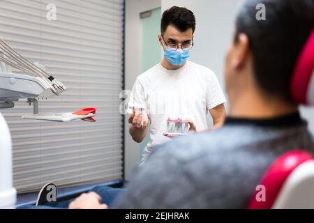 Le chirurgien dentaire avec la couronne dentaire dans les mains modèle parle au patient au sujet des prothèses. Homme au rendez-vous du dentiste Banque D'Images